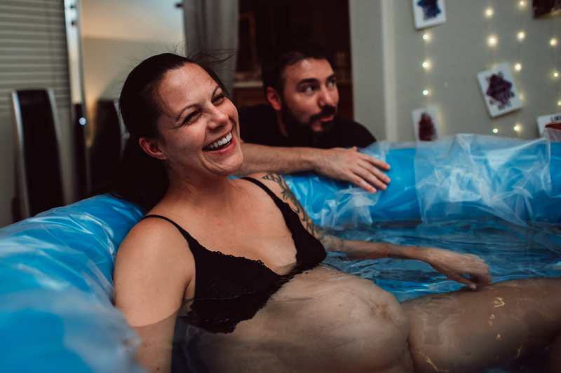 Birth Photography - woman in labor smiles as she sits in birthing pool preparing for baby's arrival